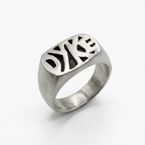 Op de afbeelding: Een geborstelde zilveren zegelring met een rechthoekige voorkant. De voorkant heeft een uitgesneden ontwerp met geometrische vormen. De ring heeft een gladde, afgeronde band en een geborstelde afwerking, wat een moderne, minimalistische uitstraling creëert.