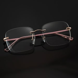 Peut inclure: Lunettes sans monture avec des verres rectangulaires. Les montures sont de couleur rose clair avec des accents dorés au niveau des charnières et du pont. Les verres sont clairs et les lunettes semblent légères.