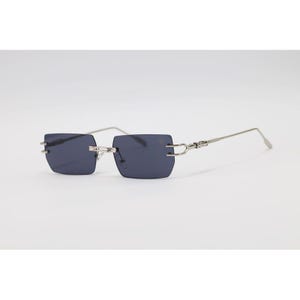 May include: Rimless sunglasses with rectangular, dark gray lenses and silver-toned metal frames. The temples are thin and straight, with decorative accents near the hinges. The sunglasses are set against a plain white background.