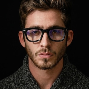 May include: A portrait of a person wearing black-framed eyeglasses. The glasses have a rectangular shape. The person has a beard and is wearing a gray sweater. The background is black.