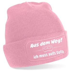 Könnte beinhalten: Hellrosa Beanie-Mütze mit rechteckigem Aufnäher. Der Aufnäher zeigt den deutschen Text "Aus dem Weg! ich muss aufs Sofa" in Weiß sowie eine Sofa-Illustration. Die Mütze ist aus Strickmaterial.