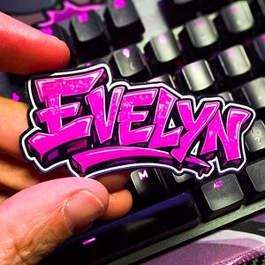 May include: A pink and purple sticker with the name "EVELYN" in a graffiti-style font. The sticker is held in front of a black keyboard with pink backlit keys. The keyboard is on a purple and black patterned surface.