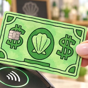May include: A rectangular payment card with a light green background and dark green outlines. The card features a central illustration of a seashell inside a circle, flanked by two dollar signs. A metallic EMV chip is embedded on the left side.