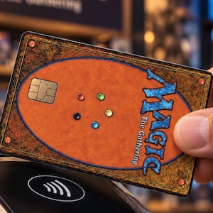 May include: A rectangular payment card featuring the Magic The Gathering logo in blue text on an orange background. The card includes a gold EMV chip on the left and five small colored circular icons in the center, with a textured brown border.