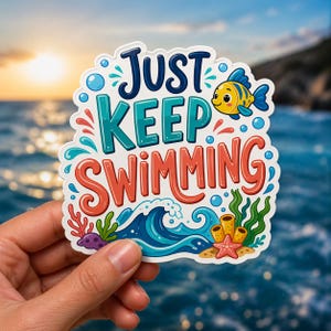 May include: A colorful sticker with the words "JUST KEEP SWIMMING" in a playful font. The design includes a cartoon fish, bubbles, waves, and underwater plants. The sticker is held in a hand against a blurred ocean background.