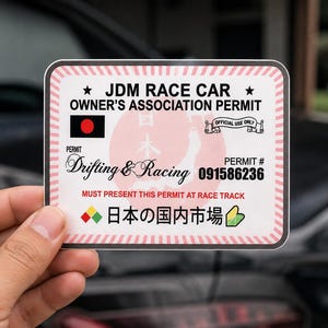 May include: A rectangular sticker with a white background and a pink striped border. It features the text JDM RACE CAR OWNER'S ASSOCIATION PERMIT, a black flag with a red circle, a map of Japan, the text Drifting & Racing, and 091586236.