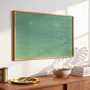 May include: A framed artwork showing a turquoise ocean scene with figures. The artwork is in a light wood frame and hangs on a white wall. A wooden cabinet with decor sits below the artwork.