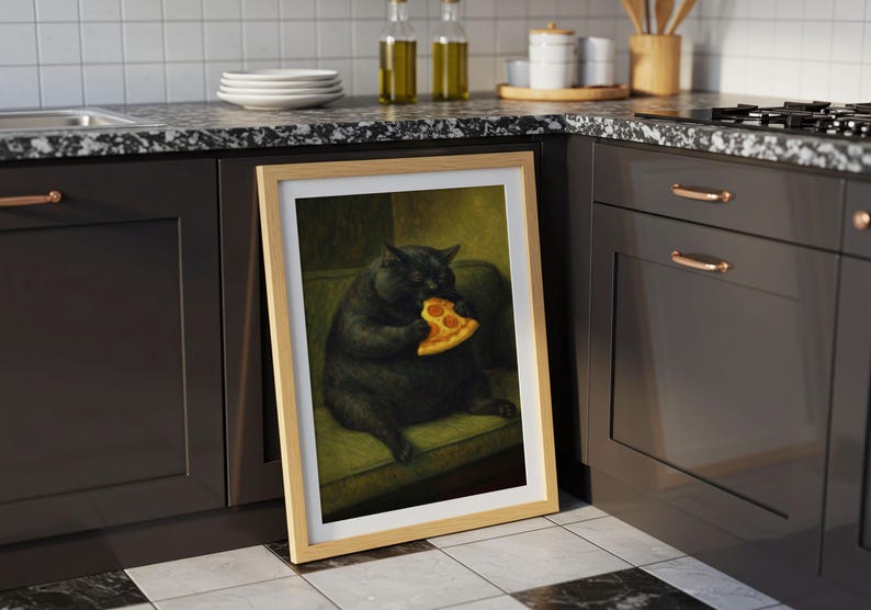 May include: A framed print featuring a black cat eating a slice of pizza. The cat is depicted sitting on a green couch. The artwork has a light wood frame and is displayed on a kitchen counter.