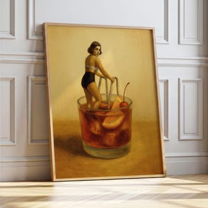 May include: A framed art print shows a woman in a swimsuit in a large cocktail glass. The glass contains a dark red liquid, orange slices, and a cherry. The artwork has a vintage, surrealist style, with a wooden frame.