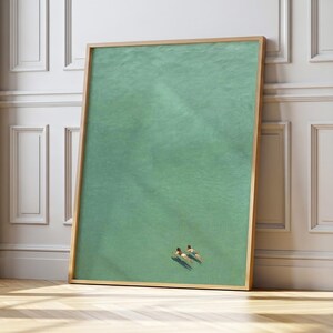 May include: A framed art print featuring an aerial view of two people swimming in turquoise water. The artwork is encased in a light-colored wooden frame, leaning against a white wall and a wooden floor.