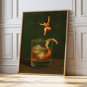 May include: A framed art print showing a cocktail glass with ice and an orange peel, with a person in a red swimsuit jumping above it. The background is dark green, and the frame is light wood.