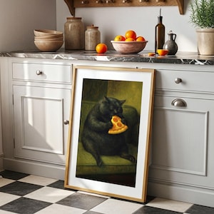 May include: A framed art print depicts a black cat seated on a green sofa, eating a slice of pizza. The artwork has a wooden frame and is leaning against a white cabinet with a marble countertop. Oranges and other kitchen items are visible.