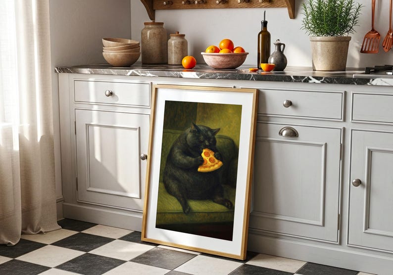 May include: A framed art print depicts a black cat seated on a green sofa, eating a slice of pizza. The artwork has a wooden frame and is leaning against a white cabinet with a marble countertop. Oranges and other kitchen items are visible.