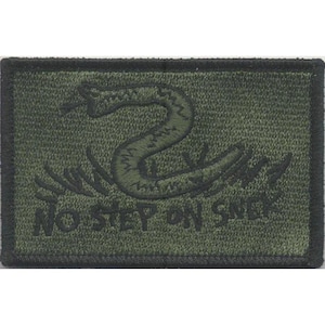 Pode incluir: Um emblema retangular bordado em verde-oliva com uma borda preta. O design apresenta um gráfico de uma cobra enrolada em verde escuro acima do texto NO STEP ON SNEK, tudo renderizado em um estilo bordado monocromático sobre tecido texturizado.