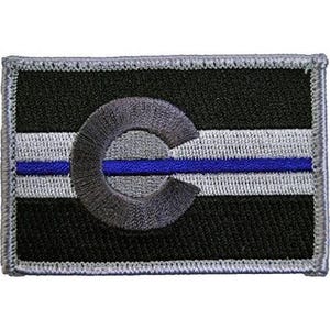 May include: A rectangular embroidered patch featuring a black background with a horizontal light grey stripe across the centre. A thin blue line runs horizontally through the stripe, and a large, stylised grey letter C is centred on the patch.
