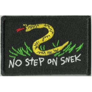 Pode incluir: Um emblema retangular preto bordado com uma cobra amarela de língua vermelha sobre grama verde. O texto NO STEP ON SNEK está bordado em letras maiúsculas brancas na parte inferior do emblema com uma borda costurada.