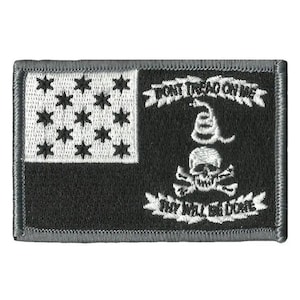 May include: A rectangular patch with a black and white design. The left side features a white rectangle with 13 black stars. The right side has a black background with a skull and crossbones, a snake, and the phrases "DON'T TREAD ON ME" and "THY WILL BE DONE."