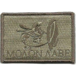 May include: A rectangular olive drab embroidered patch featuring a stylized helmet and sword graphic centered above the Greek text "ΜΟΛΩΝ ΛΑΒΕ" stitched in a matching thread color. The patch has a thick, dark brown stitched border edge.