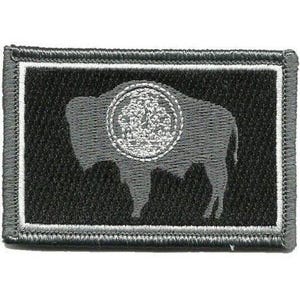 May include: A rectangular embroidered patch featuring a black background with a gray silhouette of a buffalo. The buffalo has a circular gray emblem on its side. The patch is framed by a thin white border and a thick gray stitched border edge.