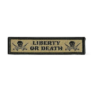 May include: A rectangular embroidered patch with a tan background and a black border. The text "LIBERTY OR DEATH" is stitched in black in the center, flanked on both sides by a black skull and crossbones icon featuring two crossed rifles.