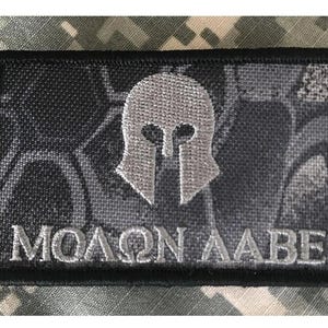May include: A rectangular patch with a black border and a gray and black background. The patch features a silver Spartan helmet design above the Greek phrase "MOLON LABE" in silver lettering. The patch is likely for military or tactical gear.