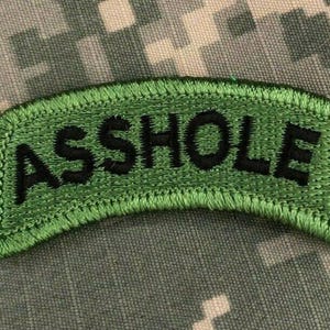 May include: Green embroidered patch with the word "ASS HOLE" in black block letters. The patch has a green border and is set against a camouflage background.