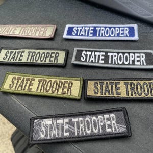May include: A collection of rectangular embroidered patches with the text "STATE TROOPER" in various color combinations. The patches include colors like tan, blue, green, and black, with white or tan lettering.