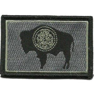 May include: A rectangular embroidered patch featuring a black silhouette of a bison on a textured gray background. The bison has a circular, textured olive green emblem on its side. The patch is framed by a thin olive green border and a thick black edge.