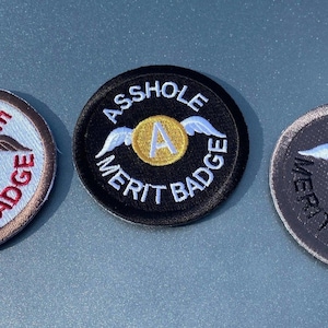 May include: Three embroidered patches with the words "ASS HOLE MERIT BADGE". Each patch is circular with a central design of a letter "A" and wings. The patches are in black, gray, and white with red and gold accents.
