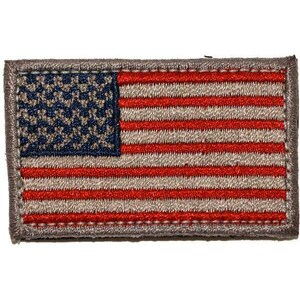 Op de afbeelding: Een rechthoekige geborduurde patch met het ontwerp van de vlag van de Verenigde Staten. De patch toont een donkerblauw kanton met een gestructureerd patroon en afwisselende horizontale strepen in rood en lichtbruin, omrand door een grijze stikrand.