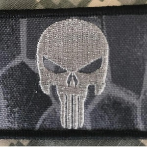 May include: A rectangular patch with a black border featuring a silver skull design. The skull has detailed features and is centered on a gray and black camouflage background. The patch is likely for clothing or gear.
