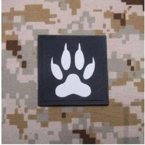May include: A square black patch featuring a white paw print graphic centered on the surface. The patch has a distinct border and a flat, matte finish, presenting a clear contrast between the dark background and the light animal footprint design.