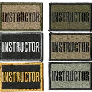May include: A collection of six rectangular embroidered patches. Each patch features the word "INSTRUCTOR" in bold, capital letters. The patches vary in color, including olive green, black, tan, and gray, with contrasting thread colors.