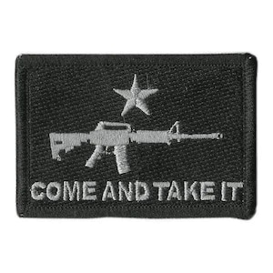 May include: A rectangular black embroidered patch featuring a grey rifle silhouette centered below a single grey star. The text COME AND TAKE IT is embroidered in grey capital letters along the bottom edge of the patch against the black background.