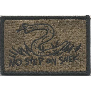 Pode incluir: Um emblema retangular bordado em verde-oliva com uma borda preta. O emblema apresenta um gráfico preto bordado de uma cobra enrolada e o texto NO STEP ON SNEK em uma fonte de estilo manuscrito. O design está centralizado no tecido.