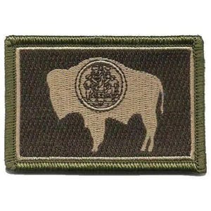 May include: A rectangular embroidered patch featuring a dark brown background with a tan silhouette of a bison. Centered on the bison is a circular emblem with intricate detailing. The patch is bordered by a thick olive green stitched edge.