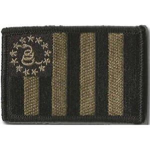 May include: A rectangular embroidered patch featuring a black and dark olive green color scheme. The design displays a stylized flag with vertical stripes and a circular arrangement of thirteen stars surrounding a coiled snake emblem on the left.