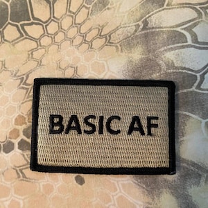 May include: A rectangular patch with the embroidered text "BASIC AF" in black on a tan background. The patch has a black border and is set against a camouflage pattern.
