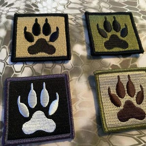 May include: Four square patches with embroidered paw prints. The patches feature a variety of colors, including tan, olive green, black, and gray, with the paw prints in contrasting colors like black and white. The patches are likely for clothing or accessories.