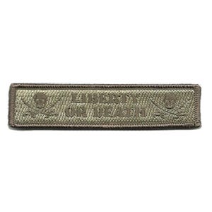 May include: A rectangular olive green embroidered patch with a dark brown border. The text LIBERTY OR DEATH is centered in the middle, flanked by two skull and crossbones icons on the left and right sides, all rendered in a monochromatic thread.