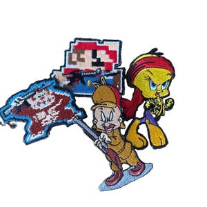 May include: A collection of embroidered patches featuring a pixelated Mario character, a pixelated brown creature holding a blue flag, a cartoon character in a tan hunting outfit with a rifle, and a yellow bird character wearing a red head wrap.