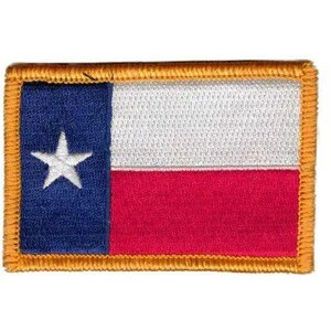 Pode incluir: Um emblema retangular bordado com o design da bandeira do estado do Texas. Possui uma faixa vertical azul à esquerda com uma estrela branca e faixas horizontais branca e vermelha à direita, tudo contornado por um fio de cor dourada.