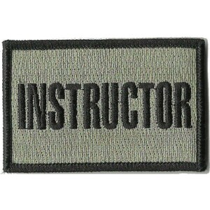 May include: A rectangular embroidered patch with a sage green background and a thick black border. The word INSTRUCTOR is stitched in bold, black, capital letters across the center of the patch, featuring a textured fabric finish throughout.