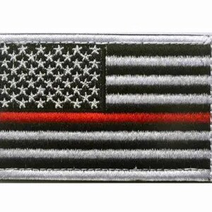 May include: Embroidered patch featuring a stylized American flag design. The flag has a black and white color scheme with a red stripe and white stars. The patch has a silver border.