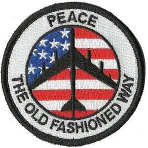 May include: Embroidered patch with a circular design. The patch features a peace symbol overlaid on a stylized American flag. The words "PEACE" and "THE OLD FASHIONED WAY" are embroidered around the peace symbol.