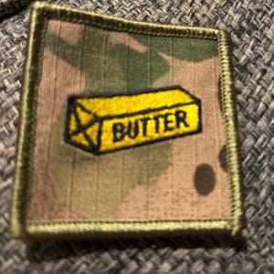 May include: Square patch with a camouflage background and a yellow rectangle with the word "BUTTER" in black. The patch has a green border.