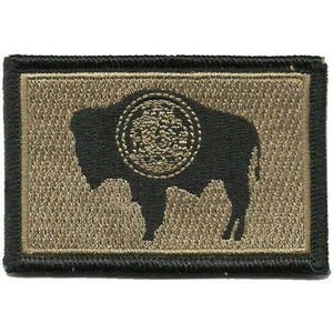 May include: A rectangular embroidered patch featuring a black silhouette of a bison on a tan background. The bison has a circular textured emblem on its side. The entire patch is framed by a thick black stitched border.