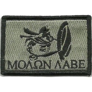 Puede incluir: Un parche rectangular bordado de color verde oliva con un borde negro. El diseño presenta un gráfico negro bordado de un casco espartano y un escudo, con el texto griego "ΜΟΛΩΝ ΛΑΒΕ" cosido en letras negras a lo largo del borde inferior.