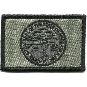 May include: A rectangular embroidered patch in olive green and black featuring the Great Seal of the State of Nebraska. The circular emblem depicts a blacksmith, a steamboat on a river, mountains, and a train, surrounded by the text The Great Seal of the State of Nebraska, March 1st, 1867.