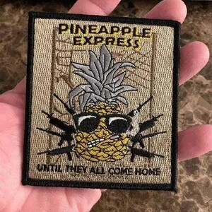 May include: Embroidered patch with the words "PINEAPPLE EXPRESS" above a pineapple wearing sunglasses and surrounded by black weapons. The text "UNTIL THEY ALL COME HOME" is below the pineapple. The patch has a black border.